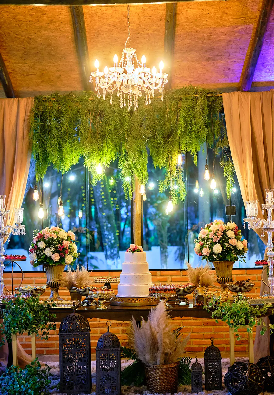 Salão à noite com lustre de cristal, cortina de folhagens e mesa do bolo